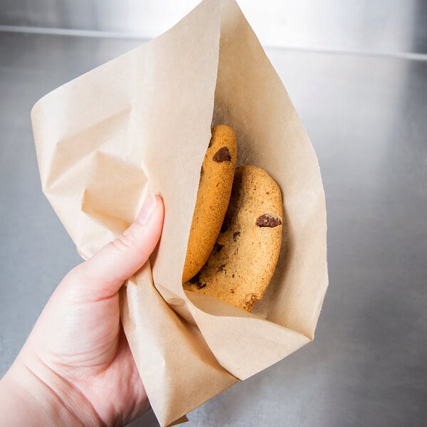A hand using a Choice 7" x 7" Natural Kraft Basket Liner to hold a paper bag with cookies.