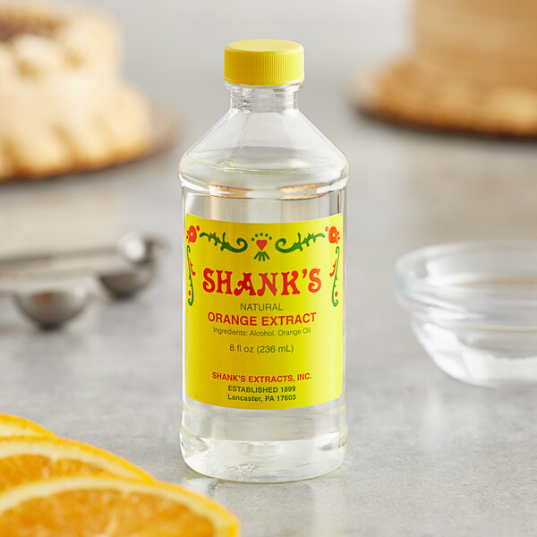 A bottle of Shank's Pure Orange Extract next to an orange slice on a counter.