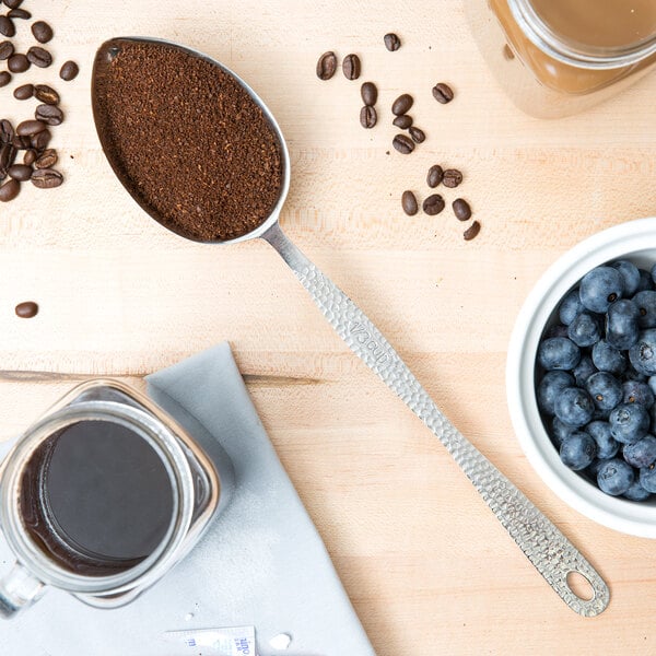A stainless steel spoon with blueberries and ground coffee next to a jar of coffee.
