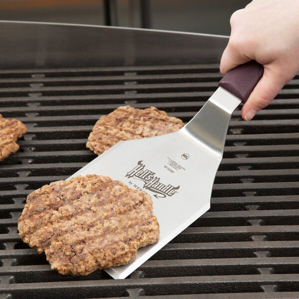 A hand using a Mercer Culinary Hell's Handle square edge turner to cut a burger on a grill.