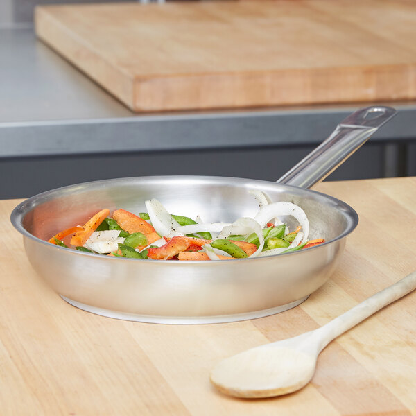 A Vollrath stainless steel fry pan with vegetables and a wooden spoon.