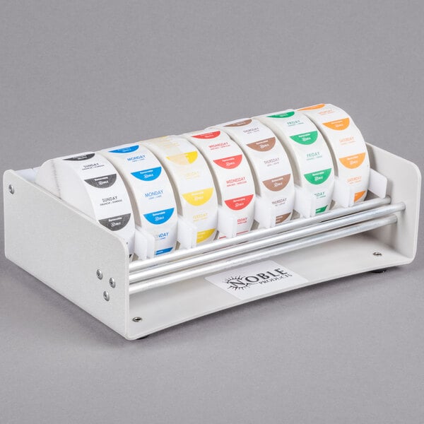 A white Noble Products dispenser rack holding seven rolls of food labeling stickers.