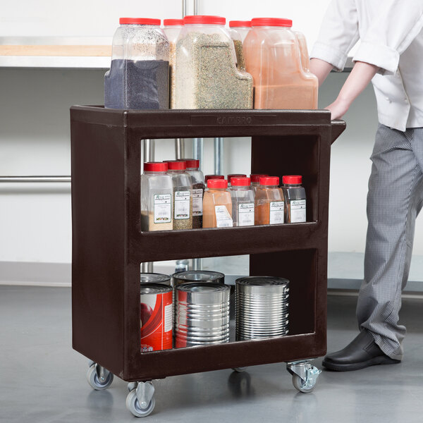 A man pushing a dark brown Cambro service cart with shelves holding food and spices.