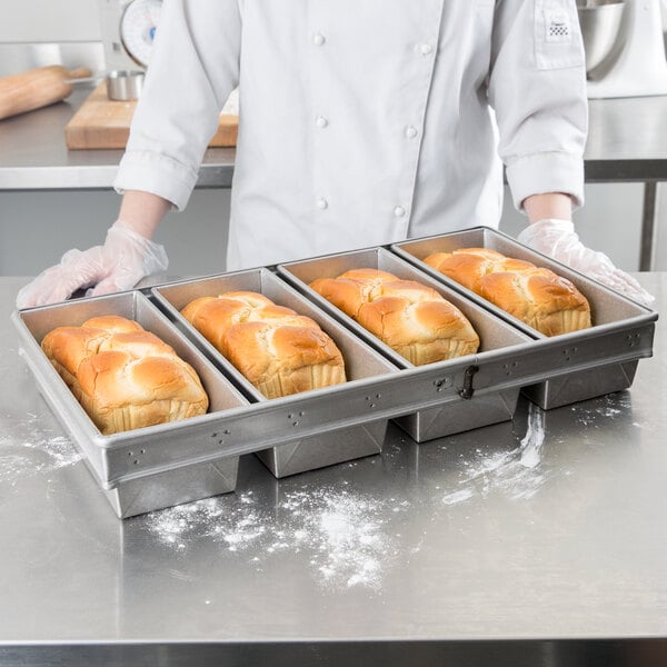 A person in a white chef's uniform holding a tray of bread loaves baked in a Chicago Metallic 4-Strap Bread Loaf Pan.