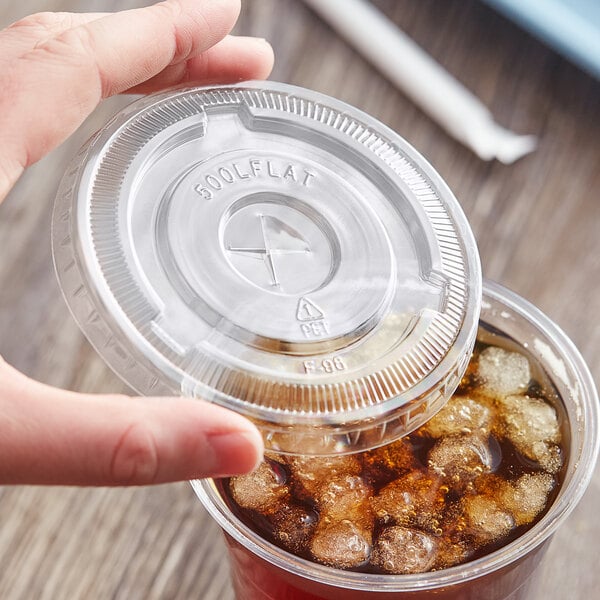A hand holding a Choice clear plastic lid with a straw slot over a plastic cup of ice.
