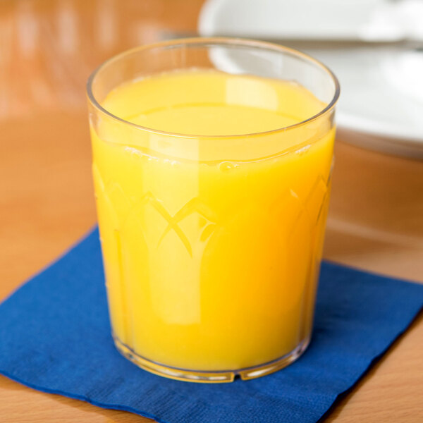 A close-up of a Dinex Fenwick clear plastic tumbler filled with orange juice on a table.
