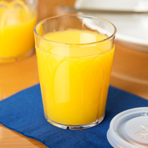 A Dinex clear plastic tumbler filled with orange juice on a table with a blue napkin.