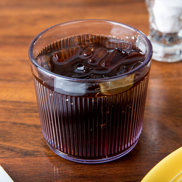 A Dinex Crystalon clear plastic tumbler filled with a brown drink and ice on a table next to a plate of food.