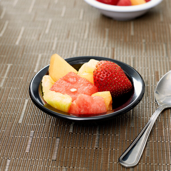 A Carlisle black rimmed melamine fruit bowl with a spoon on a table with a bowl of fruit.
