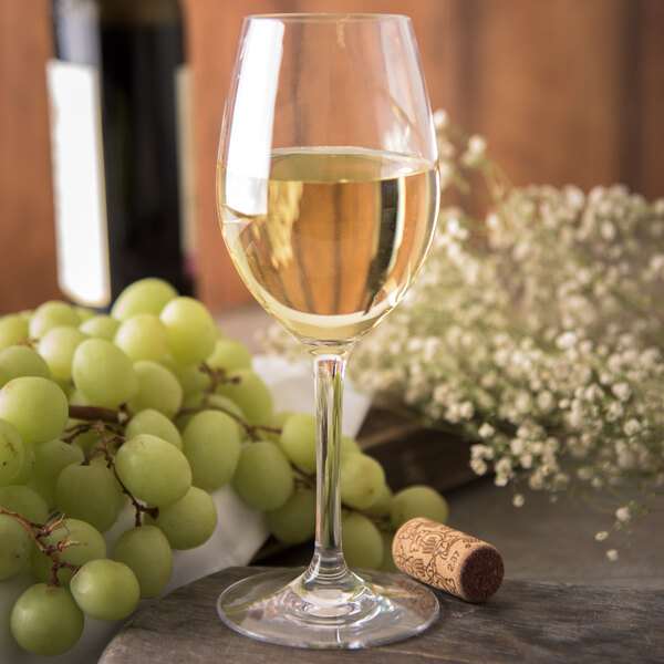 A close-up of a Carlisle plastic white wine glass full of white wine next to a bottle and grapes.