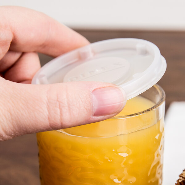 A hand placing a Dinex translucent plastic lid over a glass of orange juice.