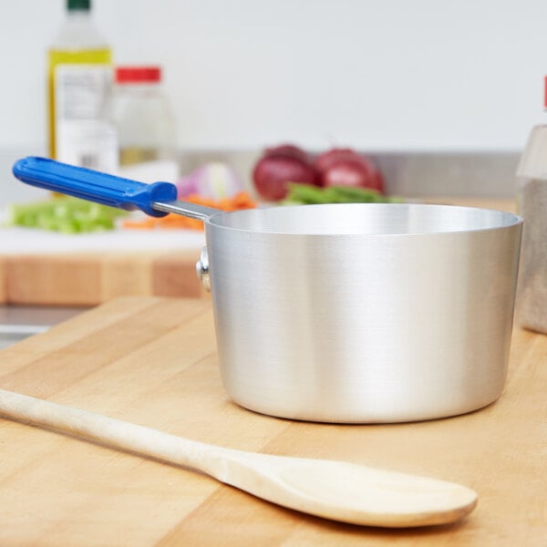 A silver Vollrath sauce pan with a blue silicone handle on a wooden cutting board.