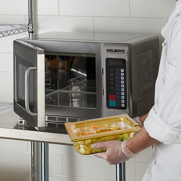A person in a white coat holding a tray of food uses a Solwave commercial microwave.