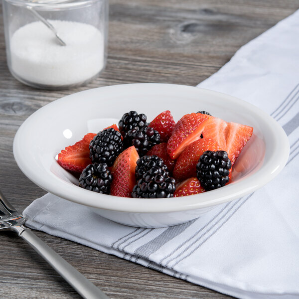 A Fiesta white china cereal bowl filled with strawberries and blackberries on a table with a glass of milk and a spoon.