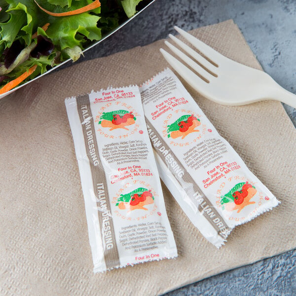 Two packets of Italian dressing on a table with a salad and fork.
