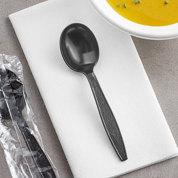 An individually wrapped black plastic spoon on a napkin next to a bowl of soup.