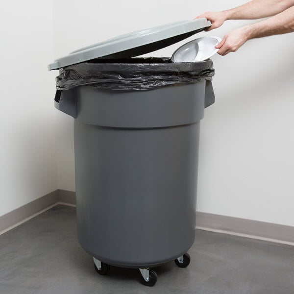 A person putting a white lid on a grey Continental Huskee trash can with a black plastic bag over it.