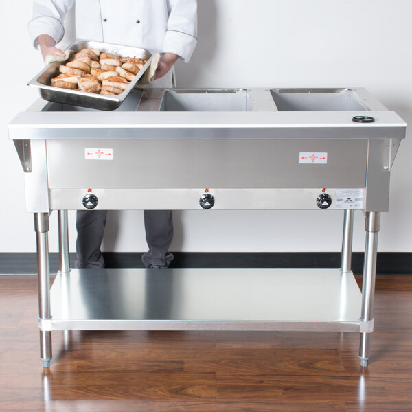 A chef holding a tray of food in front of an Advance Tabco electric steam table with undershelf.