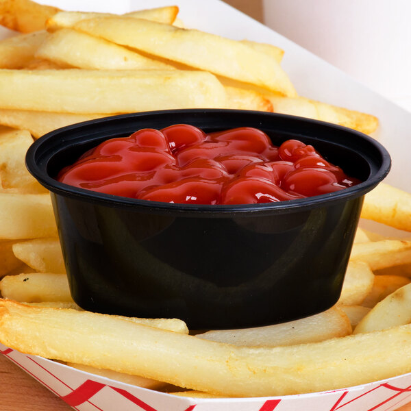 A black Newspring oval plastic souffle cup filled with french fries and ketchup.