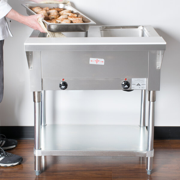 A person holding a pan in a commercial kitchen on an Advance Tabco electric hot food table.