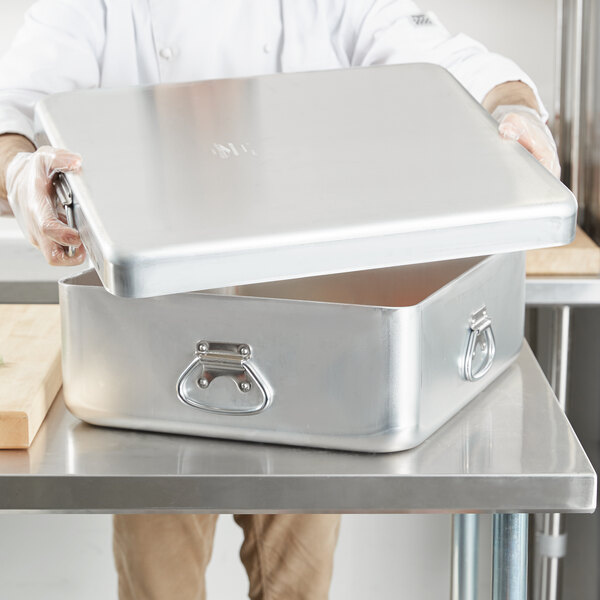 A person holding a Vollrath aluminum roasting pan with handles.