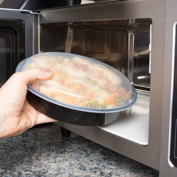 A hand holding a Pactiv black plastic container of food in a microwave.