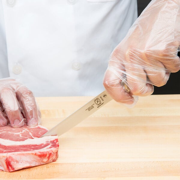 A person in a white glove using a Mercer Culinary Genesis boning knife to cut meat on a counter.
