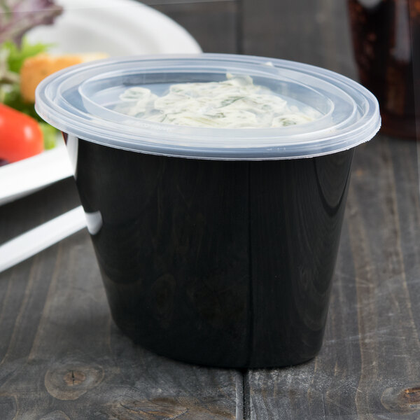 A black Newspring oval plastic souffle cup with a clear lid on a wooden surface.