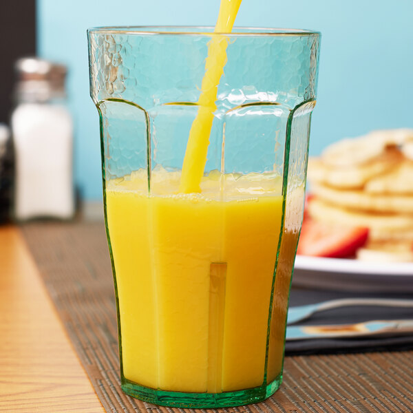 A hand pours orange juice into a Cambro Spanish Green plastic tumbler on a table.