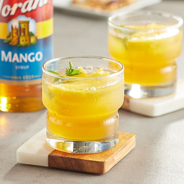 A glass of yellow liquid with a green leaf next to a bottle of Torani Mango Fruit Syrup.