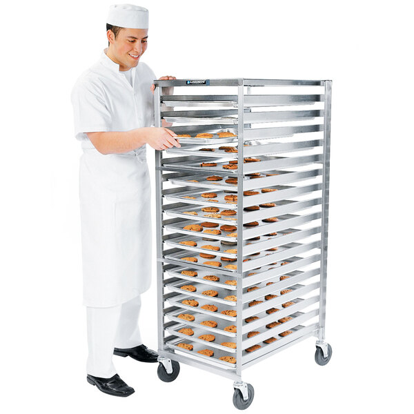 A man in a white chef uniform standing next to a Lakeside stainless steel sheet pan rack filled with trays of bread.