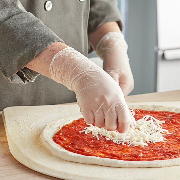 A person wearing Noble Products small powder-free clear vinyl gloves putting cheese on a pizza.