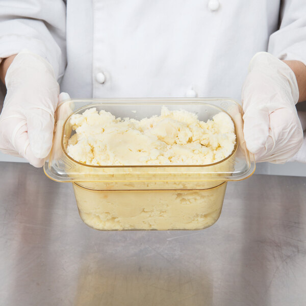 A person in white gloves holding a Cambro amber plastic food pan filled with mashed potatoes.