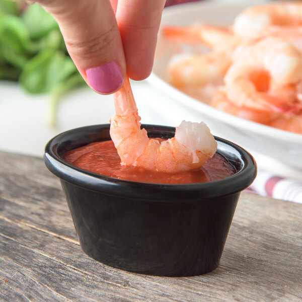 A hand holding a shrimp in a black oval ramekin of red sauce.