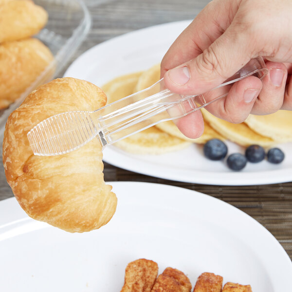 A hand using Thunder Group clear polycarbonate tongs to pick up a croissant over a plate of food.