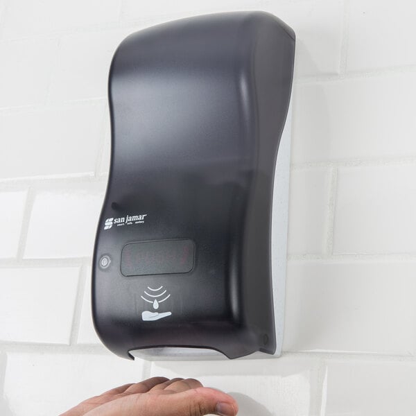 A black San Jamar Rely Pearl hybrid touchless foam soap dispenser on a wall above a hand.