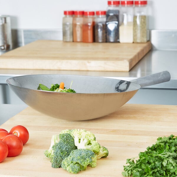 A Vollrath carbon steel wok on a cutting board with a bowl of vegetables.