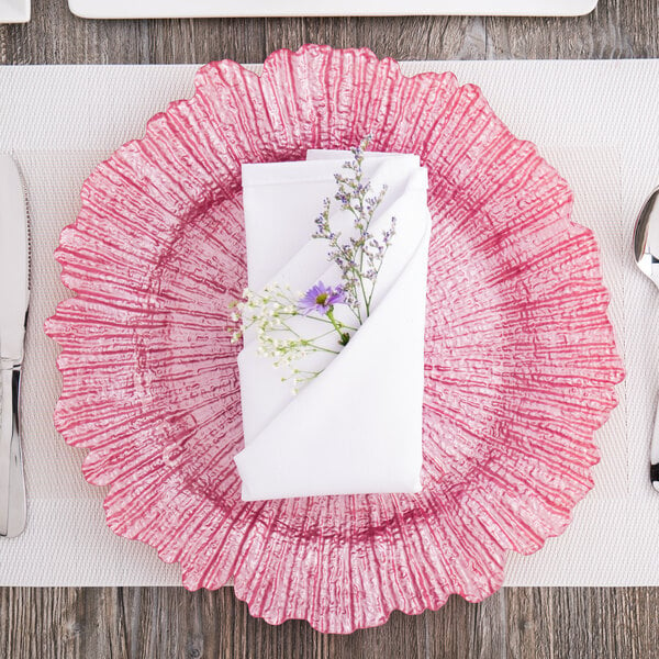 A table set with a pink Charge It by Jay glass charger plate with a napkin and flowers on it.