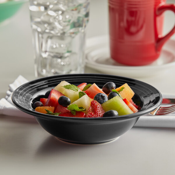A Tuxton Concentrix black china grapefruit bowl filled with fruit salad on a table with a glass of water and a blurry red coffee mug.
