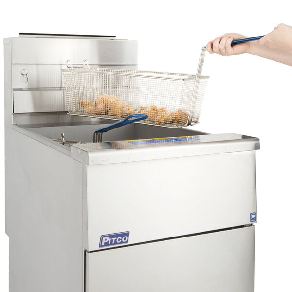 A hand holding a basket with food over a Pitco stainless steel gas fryer.