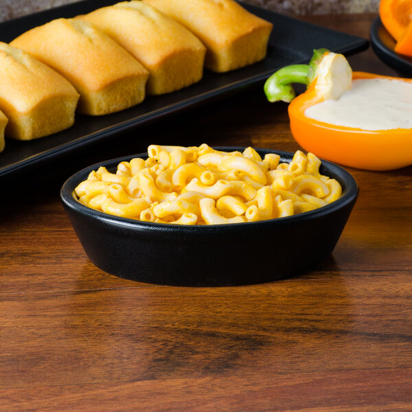 A black Hall China oval baker dish filled with macaroni and cheese on a black surface with a side of bread.