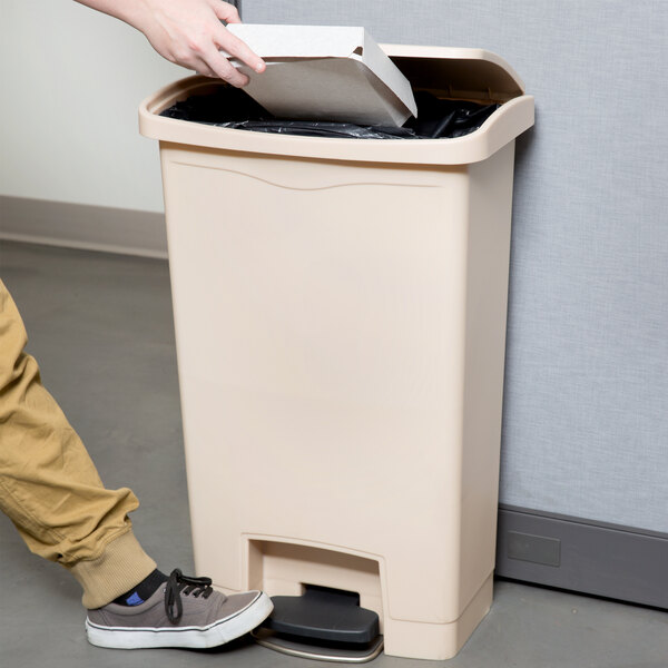 A person using their foot to open a Rubbermaid Slim Jim beige rectangular trash can lid.
