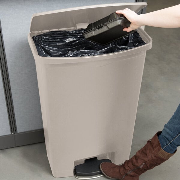 A woman uses a black plastic container to open a Rubbermaid Slim Jim beige trash can.