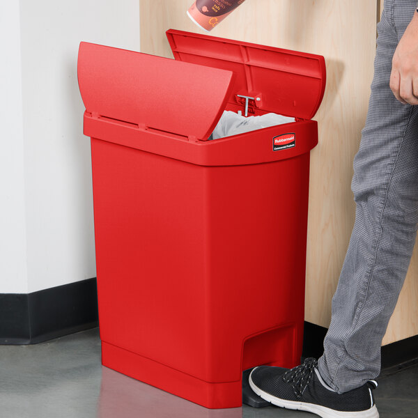 A person standing next to a red Rubbermaid Slim Jim step-on trash can with the lid open.