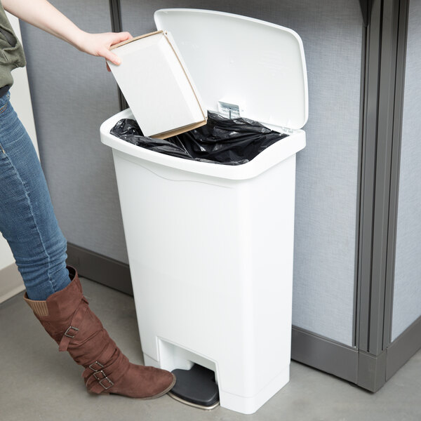 A woman using a Rubbermaid white Slim Jim front step-on trash can to put a book inside.