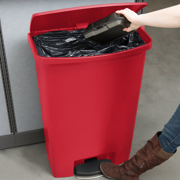 A woman using a Rubbermaid Slim Jim red front step-on trash can to throw away a black container.