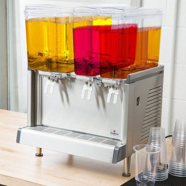 A Crathco triple bowl refrigerated beverage dispenser with different colored drinks in clear plastic cups.