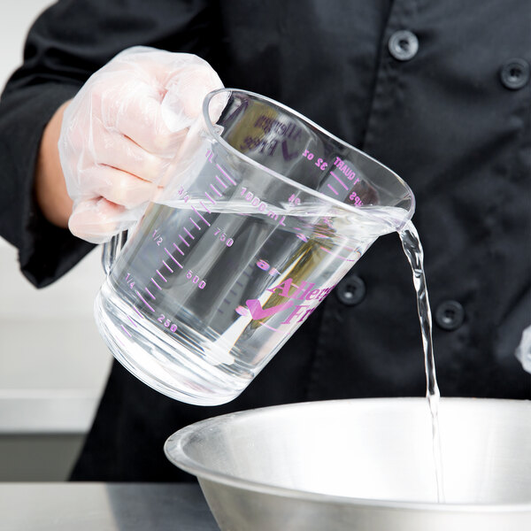 A person in a chef's uniform pouring water from a bowl into a purple Cambro measuring cup.