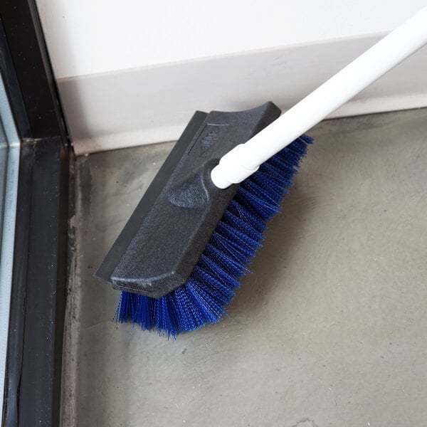 A Carlisle blue and black floor scrub brush with a squeegee on the floor cleaning a bathroom.