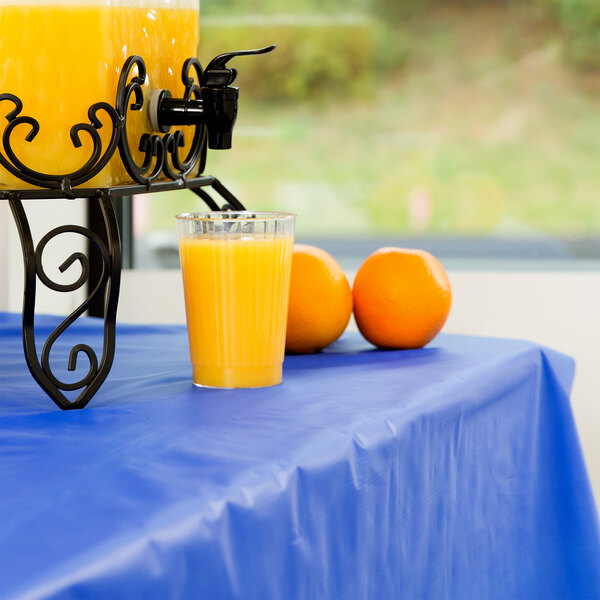 A table with a Creative Converting cobalt blue plastic table cover and a glass of orange juice on it.